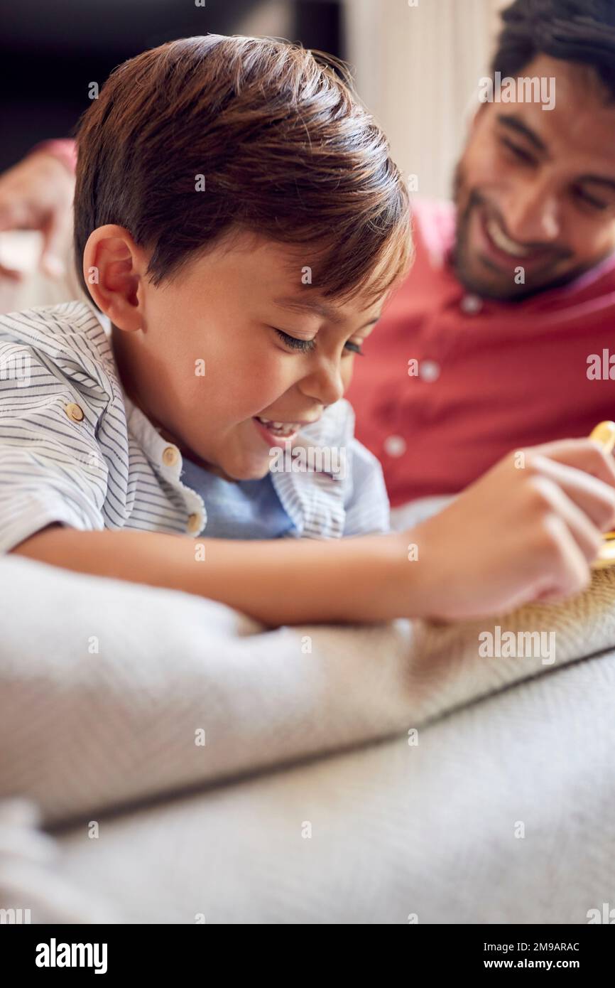 Father And Son At Home On Sofa In Lounge Playing With Digital Tablet Or Electronic Toy Together Stock Photo