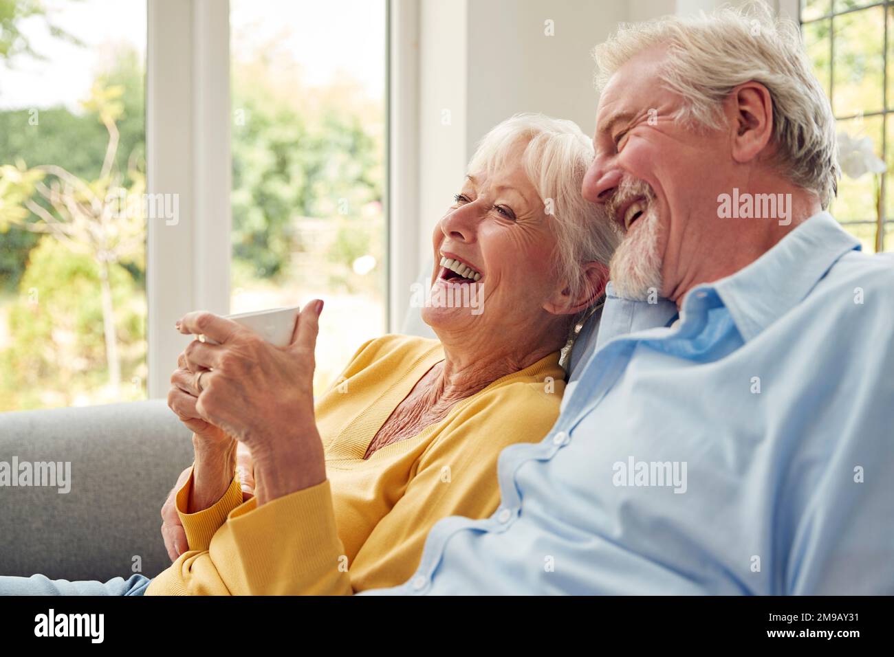 Retired Senior Couple Sitting On Sofa At Home Drinking Coffee And Watching TV Together Stock Photo