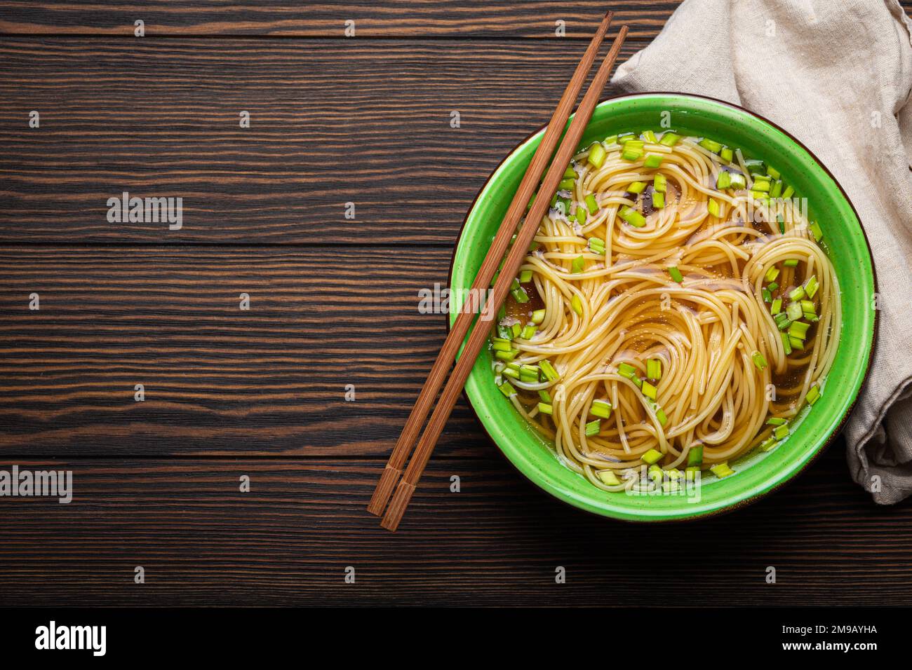 Asian noodles soup in green rustic ceramic bowl with wooden chopsticks top view on dark wooden background. Lo mein noodles with broth, green onion Stock Photo