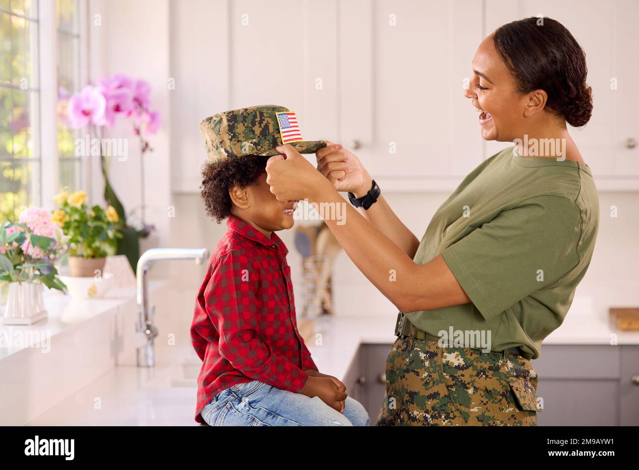 American Army Mother In Uniform Home On Leave Putting Cap On Son Sitting In Family Kitchen Stock Photo