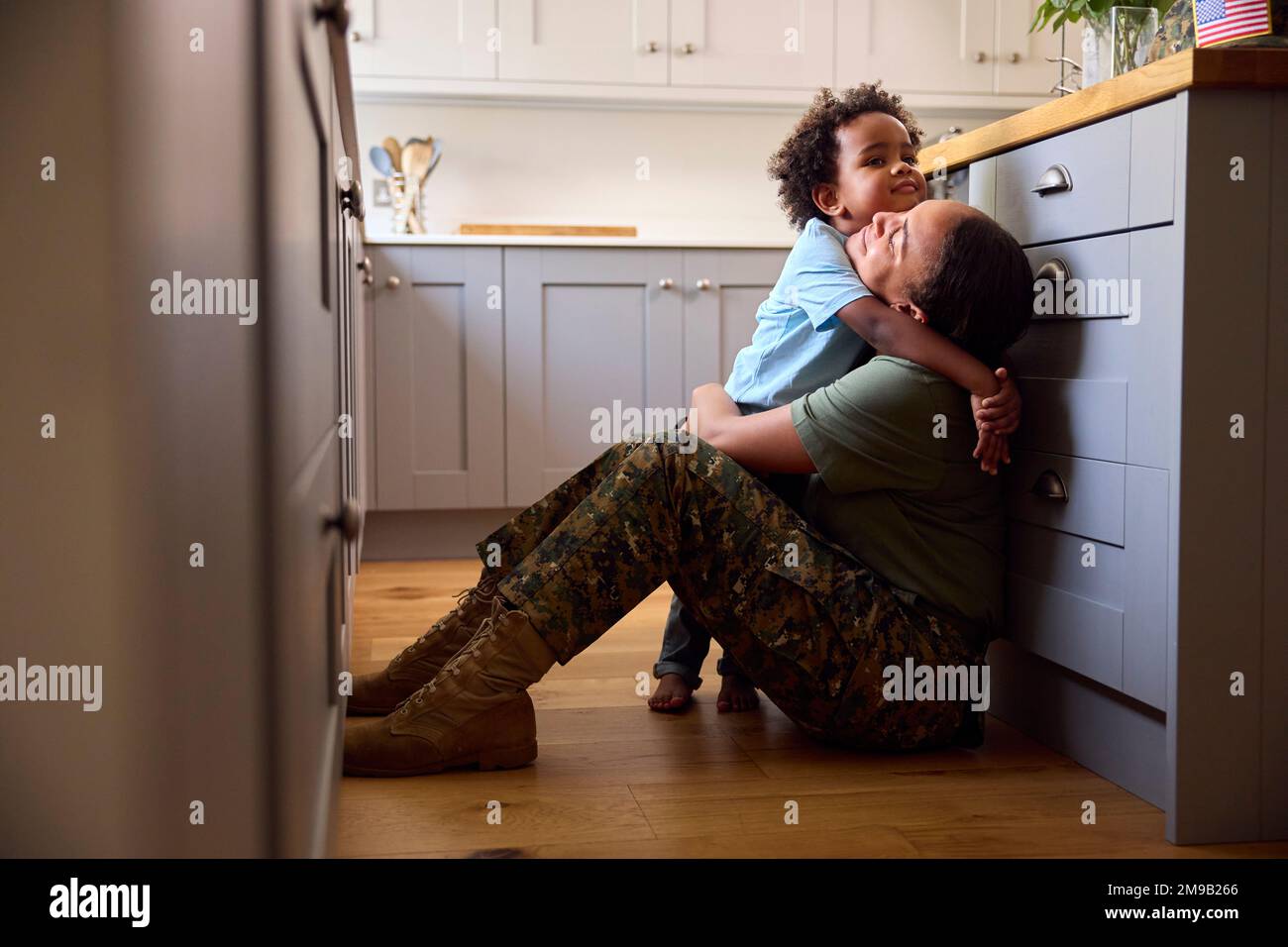 American Army Mother In Uniform Home On Leave Hugging Son Sitting On Floor In Family Kitchen Stock Photo