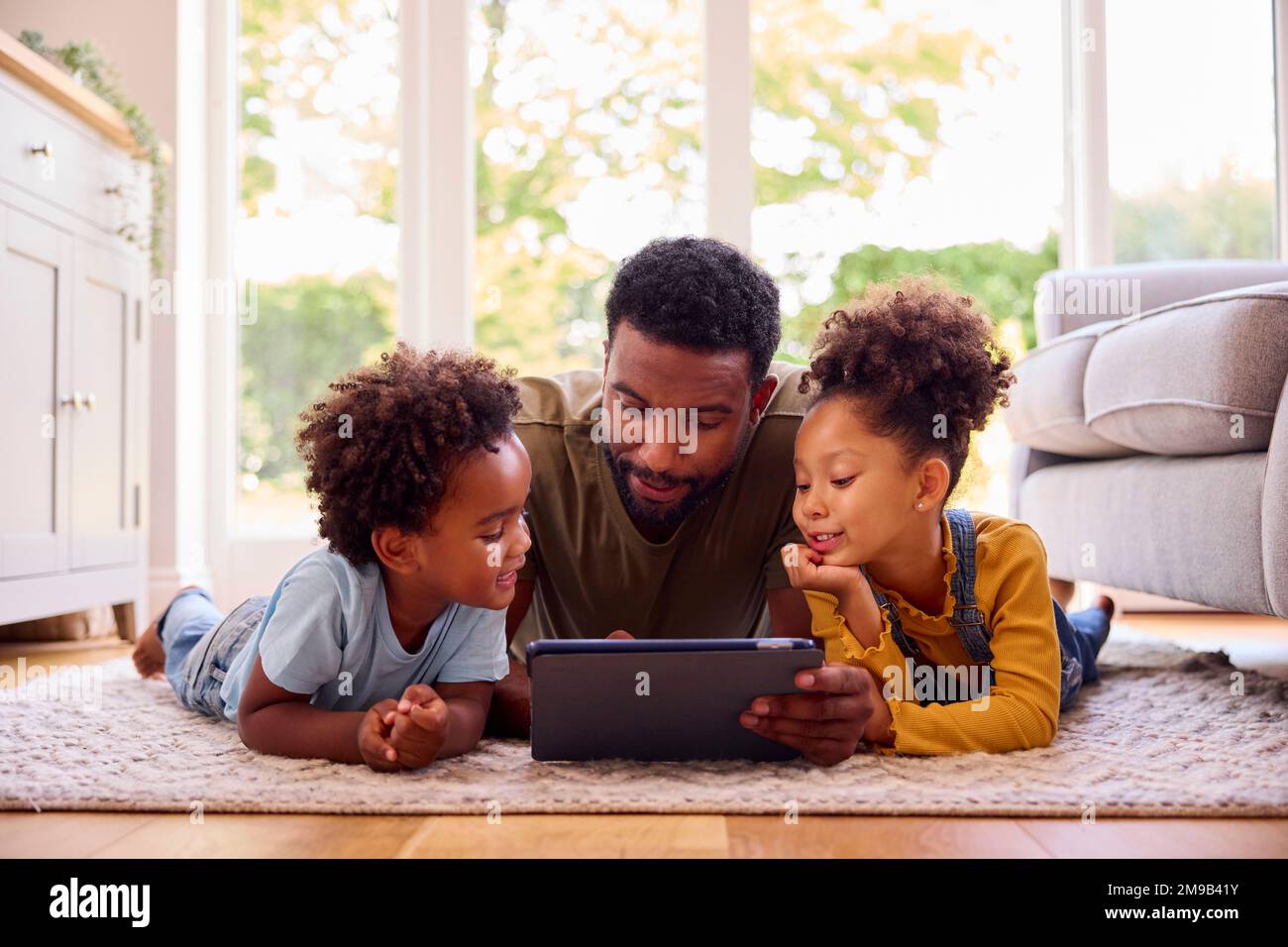 Father At Home Lying On Rug In Lounge With Children Using Digital Tablet Stock Photo