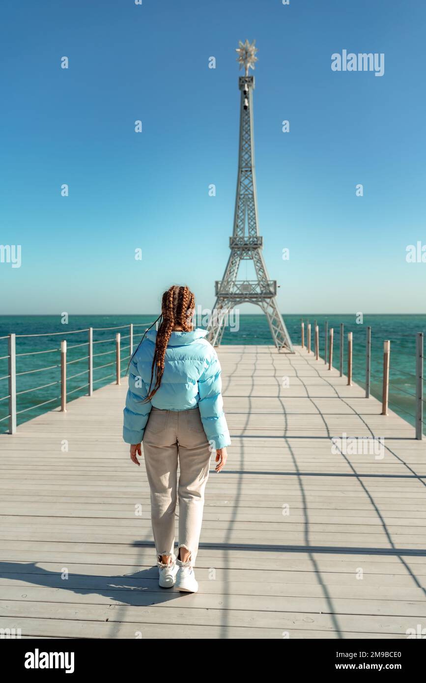 Large model of the Eiffel Tower on the beach. A woman walks along the pier towards the tower, wearing a blue jacket and white jeans. Stock Photo