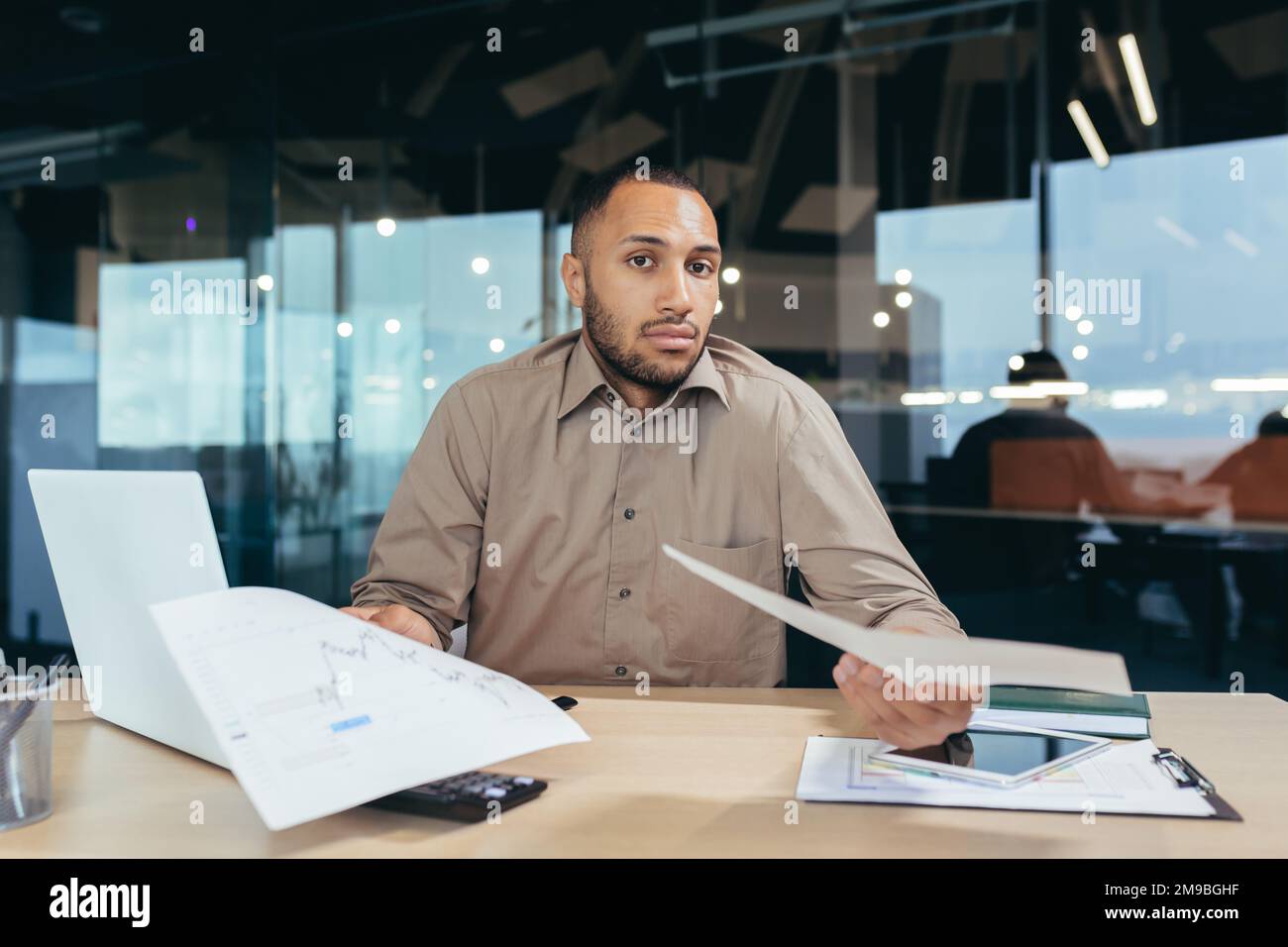 Dissatisfied businessman shows documents financial reports to camera, frustrated financier behind paper work looks at camera upset, latin american man working inside office on laptop. Stock Photo