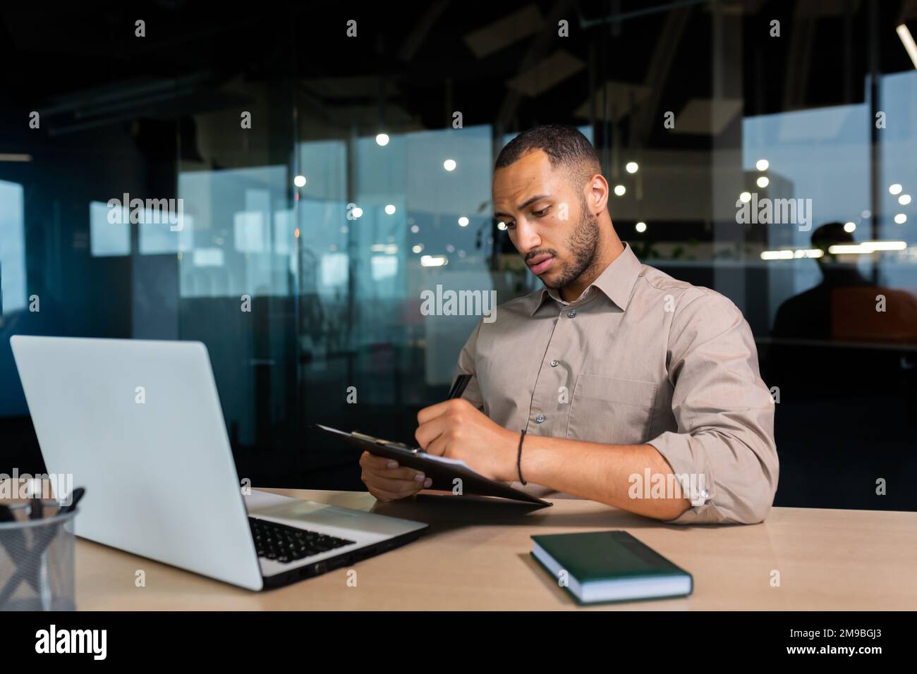 Serious concentrated businessman writing document while sitting in office using laptop for online learning, hispanic man in glasses listening to video call report at workplace. Stock Photo