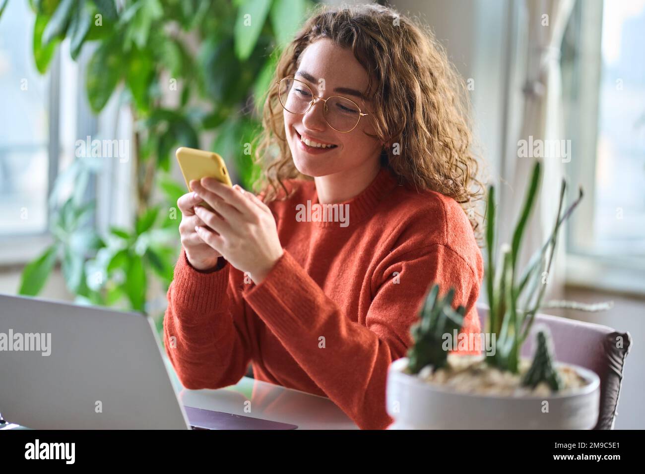 Young happy pretty woman sitting at table holding smartphone using cellphone. Stock Photo