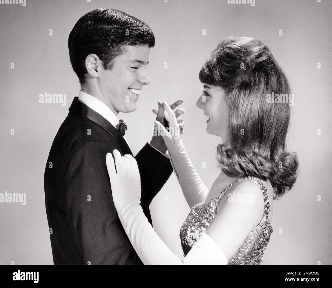 1960s TEEN COUPLE FORMAL ATTIRE DANCING ARM IN ARM AT SCHOOL PROM  - d5536 HAR001 HARS STYLE YOUNG ADULT TEAMWORK PLEASED JOY LIFESTYLE CELEBRATION FEMALES STUDIO SHOT HEALTHINESS COPY SPACE FRIENDSHIP HALF-LENGTH PERSONS MALES TEENAGE GIRL TEENAGE BOY ENTERTAINMENT CONFIDENCE B&W DATING SCHOOLS TUX HAPPINESS CHEERFUL ARM IN ARM HAIRSTYLE ATTRACTION HIGH SCHOOL SMILES HIGH SCHOOLS CONNECTION COURTSHIP JOYFUL STYLISH TEENAGED ATTIRE SEQUINS COOPERATION JUVENILES LONG WHITE GLOVES SOCIAL ACTIVITY TOGETHERNESS YOUNG ADULT MAN YOUNG ADULT WOMAN BIG HAIR BLACK AND WHITE CAUCASIAN ETHNICITY COURTING Stock Photo