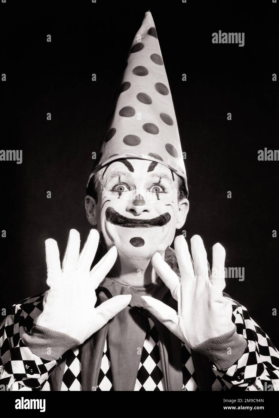 1950s 1960s SMILING FUNNY FACE CLOWN WEARING POLKA DOT DUNCE CAP RAISED HANDS IN WHITE GLOVES  - c3085 DEB001 HARS RAISED PLEASED JOY LIFESTYLE JOBS STUDIO SHOT COPY SPACE PERSONS CHARACTER MALES B&W DUNCE WIDE CLOWNS EYE CONTACT BIZARRE DOT SKILL BUG-EYED HUMOROUS OCCUPATION HAPPINESS SKILLS WEIRD HEAD AND SHOULDERS CHEERFUL PERFORMER POLKA GROTESQUE EXCITEMENT MAKE UP ZANY COMICAL UNCONVENTIONAL ENTERTAINER OCCUPATIONS SMILES COULROPHOBIA CONCEPTUAL COMEDY JESTER JOYFUL WACKY DEB001 IDIOSYNCRATIC WIDE-EYED AMUSING CREATIVITY ECCENTRIC STARTLED YOUNG ADULT MAN BLACK AND WHITE Stock Photo
