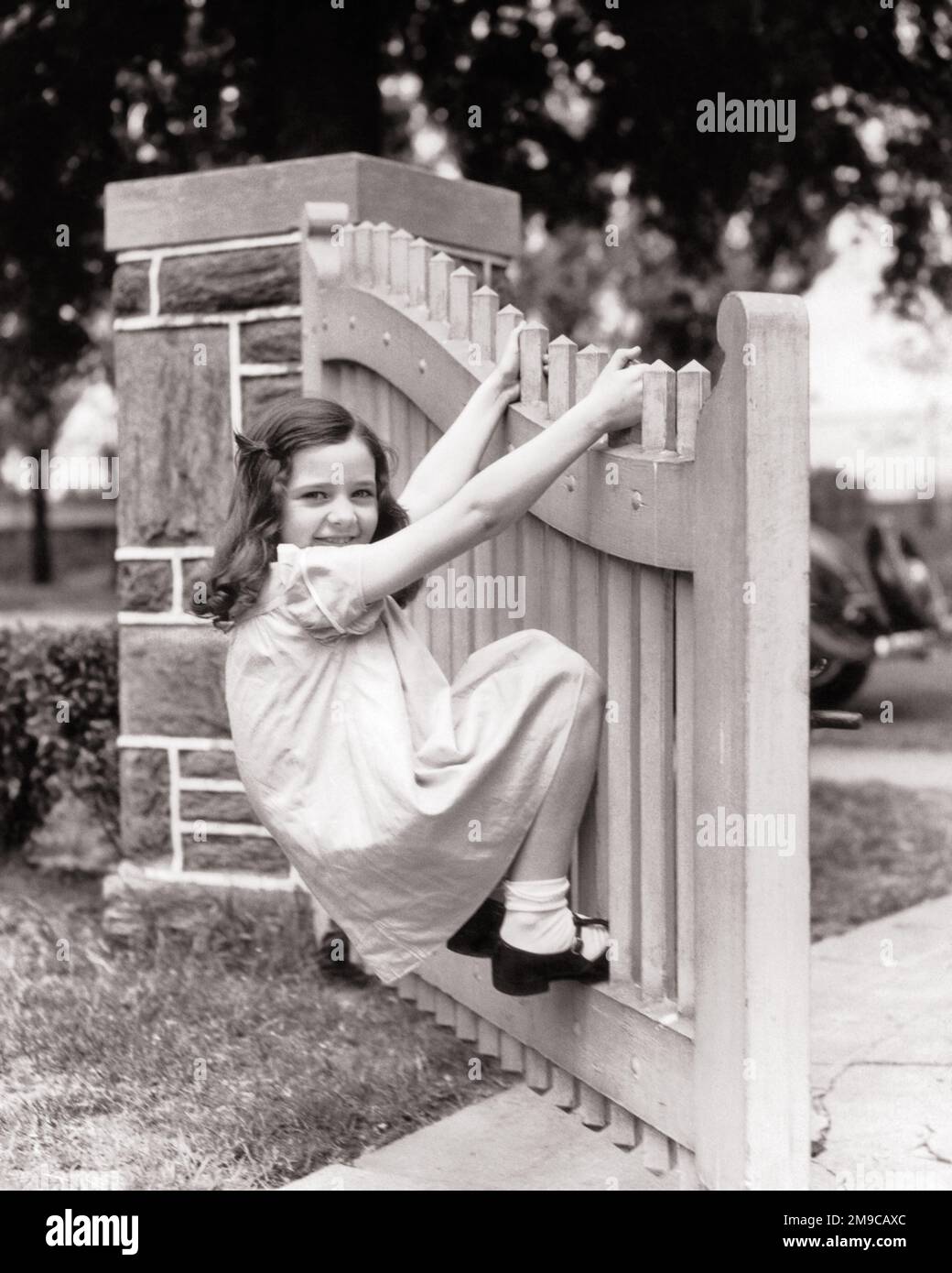 1930s SMILING BRUNETTE GIRL IN COTTON DRESS MARY JANE SHOES LOOKING AT CAMERA HANGING SWINGING ON PICKET FENCE GARDEN GATE - j547 HAR001 HARS B&W EYE CONTACT BRUNETTE GATE TEMPTATION HAPPINESS CHEERFUL ADVENTURE DISCOVERY LEISURE STRENGTH EXCITEMENT RECREATION INNOVATION PRIDE IN ON OPPORTUNITY SMILES SWINGING TOMBOY CONCEPTUAL IMAGINATION JOYFUL PICKET FENCE STYLISH COTTON DRESS GROWTH JUVENILES MARY JANE SHOES MISBEHAVING BLACK AND WHITE CAUCASIAN ETHNICITY HAR001 OLD FASHIONED Stock Photo
