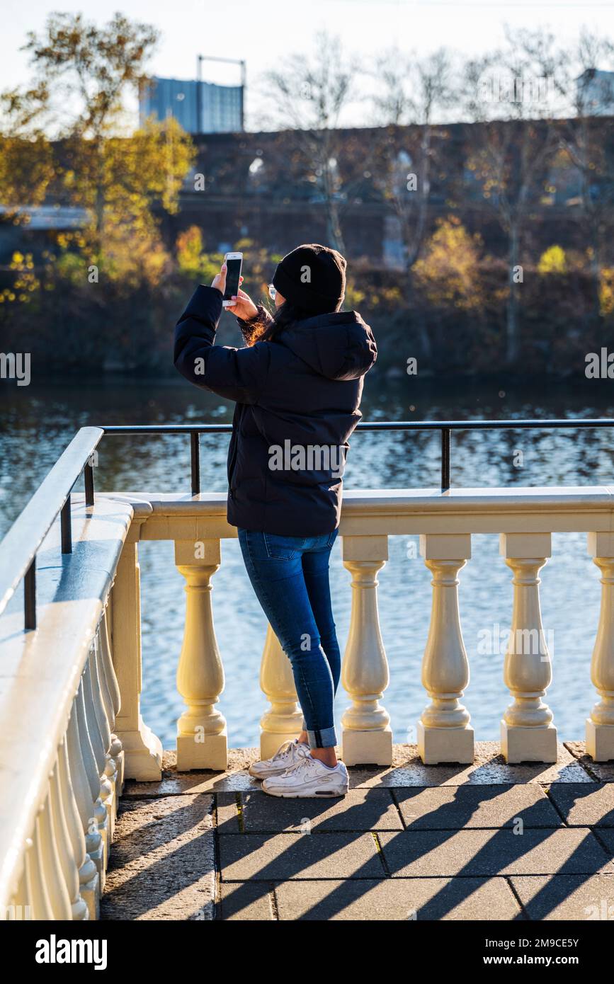 Young woman using iPhone to photograph along sunlit stone path; Fairmount Water Works; Schuylkill River; Philadelphia; Pennsylvania; USA Stock Photo