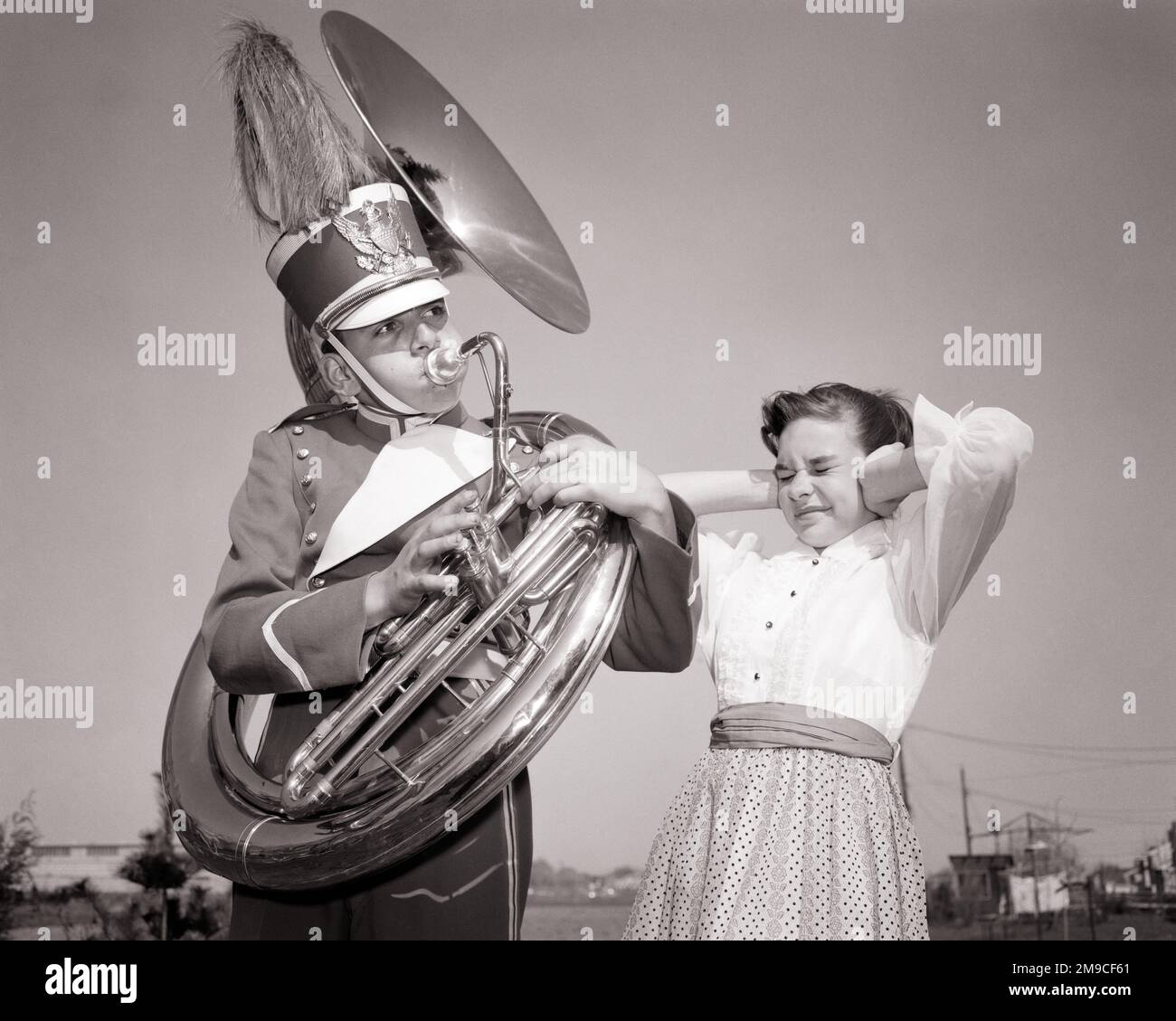 1950s TEEN BOY PLAYING TUBA LOUDLY WEARING BAND UNIFORM AND HAT YOUNG GIRL HOLDING HER HANDS OVER HER EARS FROM THE NOISE  - m2305 DEB001 HARS COMMUNICATION FRIEND COMIC NOISE LIFESTYLE SOUND SATISFACTION MUSICIAN ANNOYED FEMALES BROTHERS COPY SPACE FRIENDSHIP HALF-LENGTH INSPIRATION MALES TEENAGE GIRL TEENAGE BOY BRASS SIBLINGS SISTERS B&W HUMOROUS AND EXCITEMENT LOUD LOW ANGLE RECREATION COMICAL PRIDE SIBLING MUSICAL INSTRUMENT TUBA CONNECTION CONCEPTUAL COMEDY FRIENDLY SUPPORT DEB001 ANNOYANCE GROWTH JUVENILES LOUDLY NOISES PRE-TEEN PRE-TEEN BOY PRE-TEEN GIRL TOGETHERNESS YOUNGSTER Stock Photo