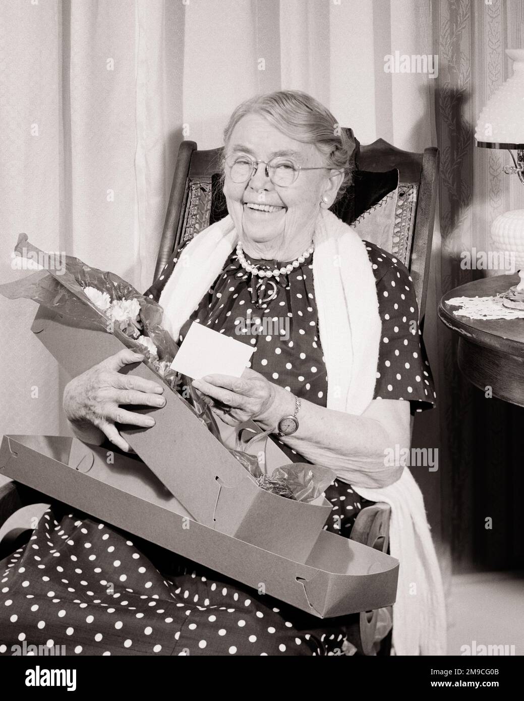 1960s SMILING GRANDMOTHER WEARING POLKA DOT DRESS SITTING IN ROCKING CHAIR HOLDING A BOX OF FRESH FLOWERS FOR MOTHER’S DAY - m7108 CRS001 HARS GRANDPARENTS PLEASED JOY LIFESTYLE CELEBRATION ELDER FEMALES GRANDPARENT HOME LIFE COPY SPACE HALF-LENGTH LADIES PERSONS SENIOR ADULT B&W EYE CONTACT SENIOR WOMAN DOT FRESH HAPPINESS OLD AGE WELLNESS OLDSTERS CHEERFUL OLDSTER POLKA MOTHER'S DAY PRIDE GRANDMOTHERS SMILES ELDERS POLKA DOT JOYFUL REMEMBERED ELDERLY WOMAN MOMS MOTHER'S BLACK AND WHITE CAUCASIAN ETHNICITY GRANDMA OLD FASHIONED REMEMBRANCE Stock Photo