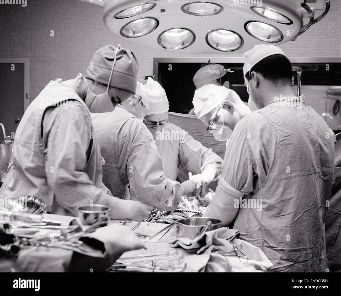 1960s DOCTORS SURGEONS NURSES IN OR STERILE CAPPED MASKED AND GOWNED PERFORMING A SURGERY UNDER HOSPITAL LIGHTING FIXTURE - m7817 HAR001 HARS OPERATING PROVIDER PROVIDERS PRACTITIONERS STERILE HEALING AND SURGICAL CAREERS PHYSICIANS SURGEONS LABOR OPERATING ROOM HEALTH CARE CAPPED EMPLOYMENT OCCUPATIONS SURGEON KCAU HEALER HOSPITALS OPERATION PHYSICIAN PRACTITIONER FACILITY INFRASTRUCTURE EMPLOYEE FACILITIES MASKED PROFESSIONALS BLACK AND WHITE CAUCASIAN ETHNICITY FIXTURE HAR001 LABORING OLD FASHIONED Stock Photo