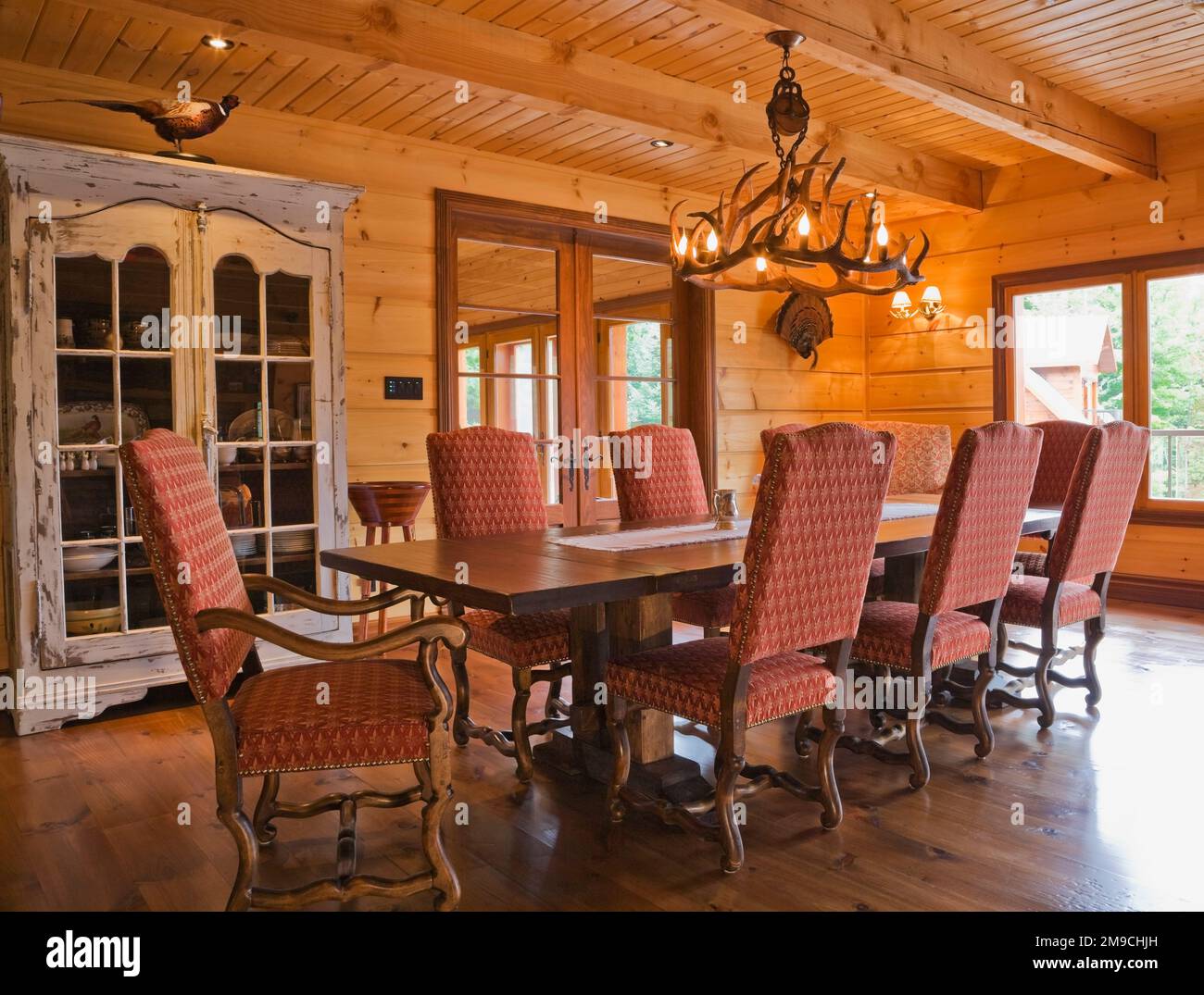 Wooden dining table with red and grey upholstered high back chairs in dining room on 1st floor inside flat log profile and timber home. Stock Photo