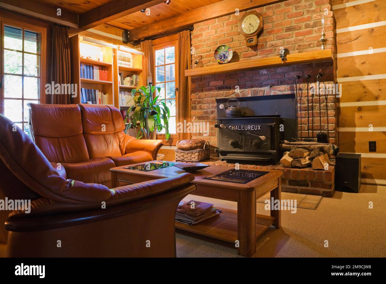 Brown leather sofas, wooden coffee table with glass tops in front of red brick wood burning stove in living room inside Canadiana style log home. Stock Photo