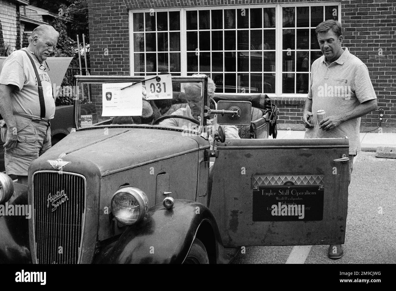 Three men gathered around a weathered vintage Austin Seven automobile at a car show in Essex, Massachusetts. The image was captured on black and white Stock Photo