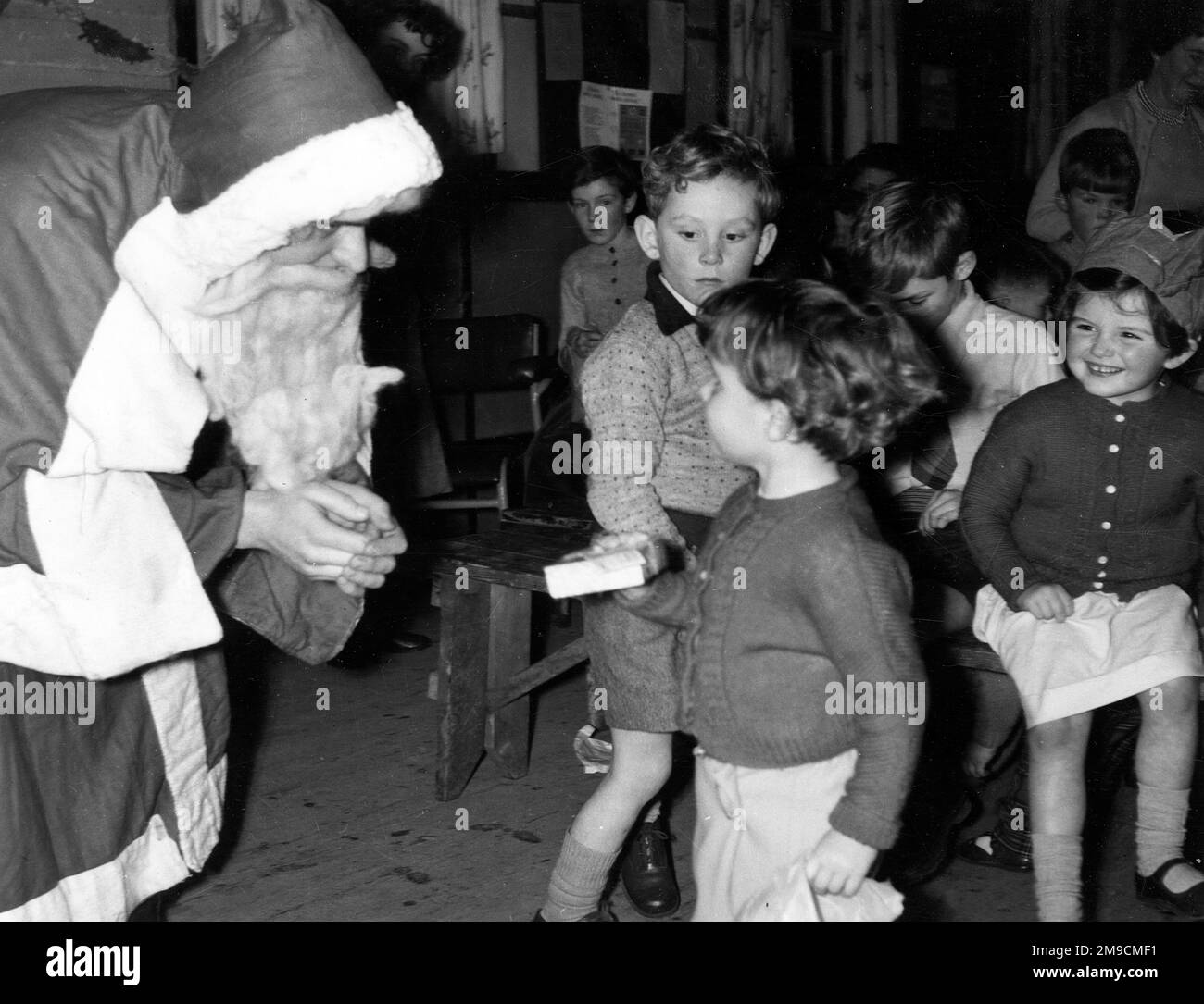 Girls and boys wait to see Father Christmas who is handing out gifts; he talks intently with one little girl in a cardigan Stock Photo