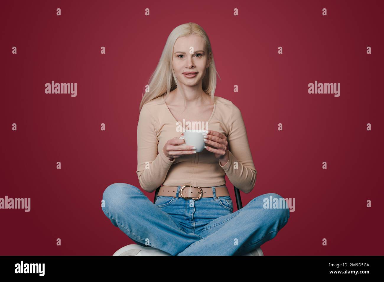 Beautiful positive woman sitting on chair holding cup of coffee and smiling at camera isolated over crimson background. Attractive beautiful girl Stock Photo