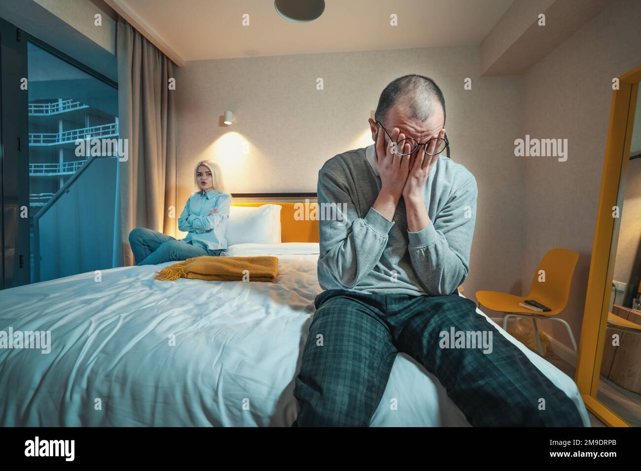 Quarrel of married couple. Man sits on edge of bed and holds his head with hands. Wife or angry girlfriend sitting cross-armed at other end of bed. Problems in relationships. Stock Photo