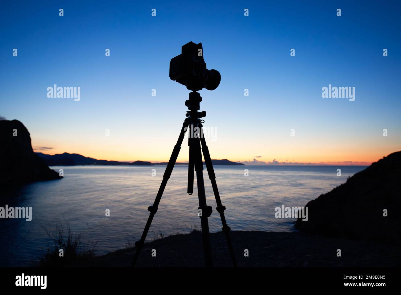 Photo camera silhouette on tripod at rocky beach with beautiful sunset in blue sea on seascape background.  Film analog photography Stock Photo