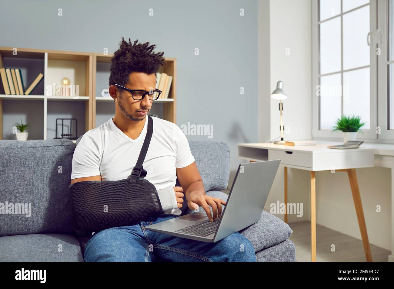 Man with broken wrist in arm sling sitting on sofa at home and working on laptop computer Stock Photo