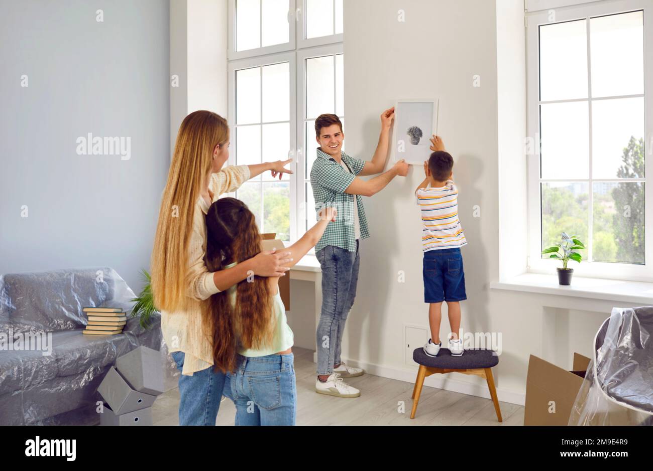 Joyful man and his little son together hang picture on wall under guidance of woman and daughter. Stock Photo