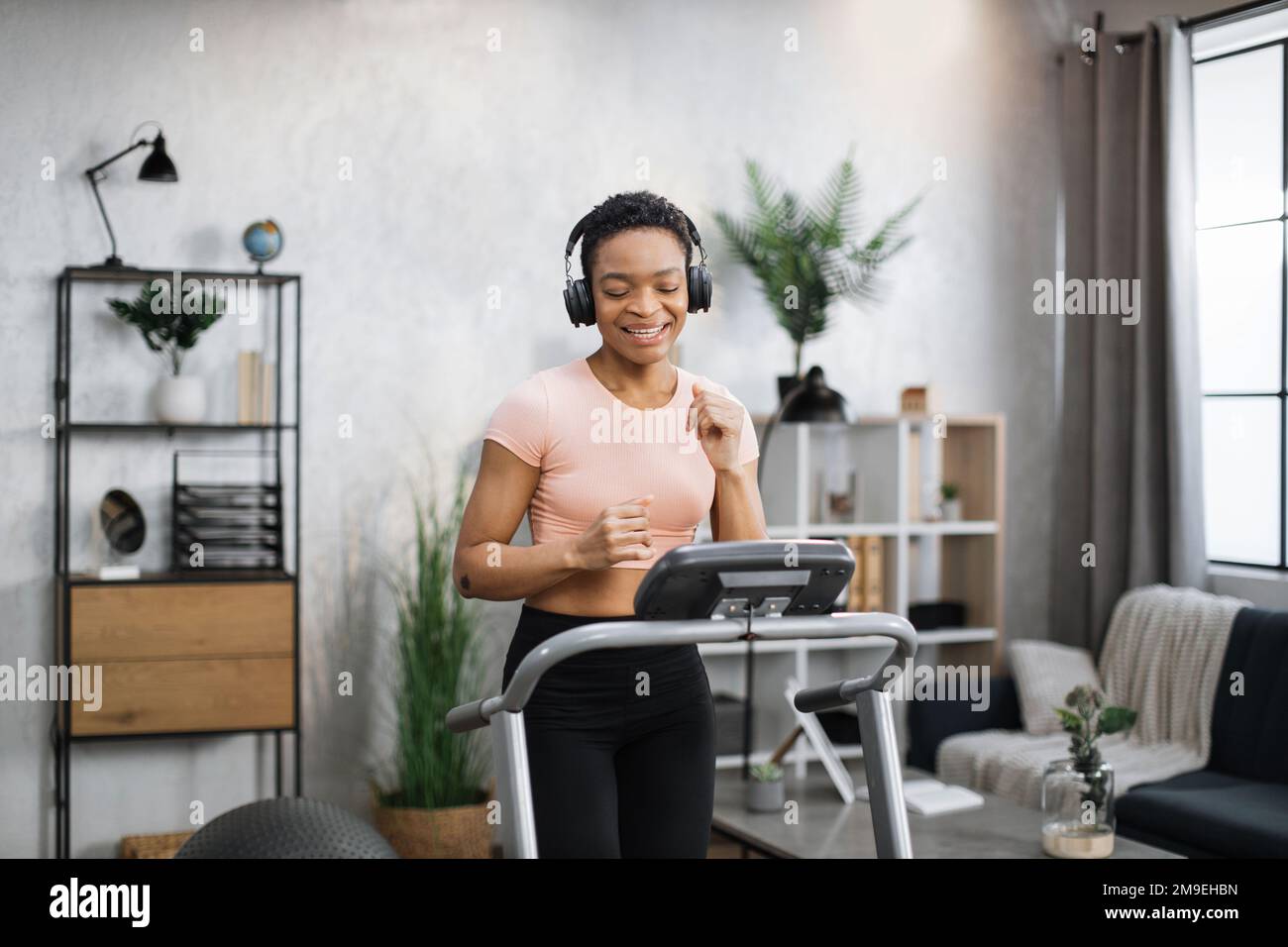 Smiling attractive young sports african woman doing fitness exercise, running on treadmill and listening music. Athletic and muscular female having actively workout indoor, at modern apartment. Stock Photo