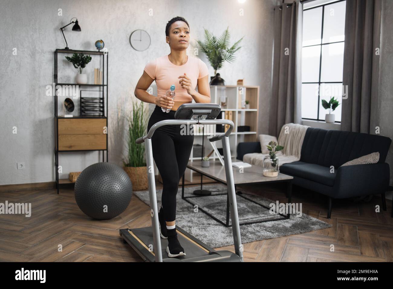 https://c8.alamy.com/comp/2M9EHXA/portrait-of-active-and-dynamic-young-charming-thirsty-african-woman-holding-water-bottle-doing-sport-fitness-at-home-running-on-treadmill-indoor-tone-your-body-perfect-shape-improving-endurance-2M9EHXA.jpg