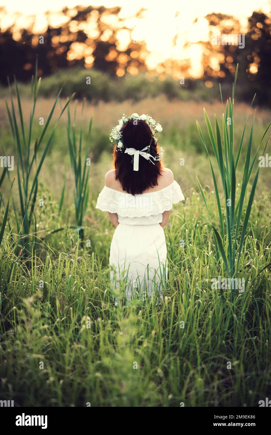 girl in a white dress with a floral wreath stands back in the field in ...