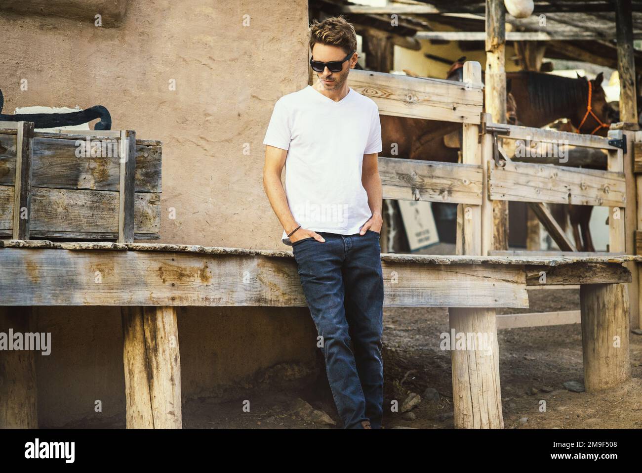 Young handsome bearded man is staying on a western city background surrounded by wooden fence. Guy is wearing a white empty t-shirt without logo. Hori Stock Photo