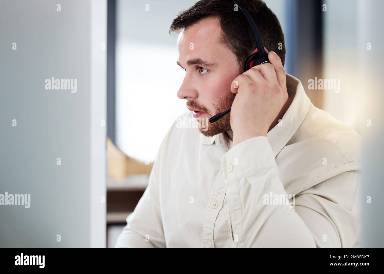 Could you repeat that. a handsome young call centre agent sitting alone in his office and looking contemplative while using his computer. Stock Photo