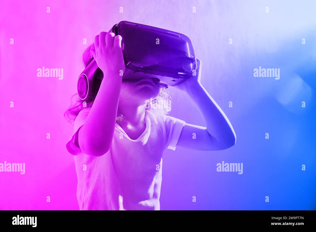 Cute curly-haired girl looks interested in VR glasses. Neon light on the background with copy space Stock Photo