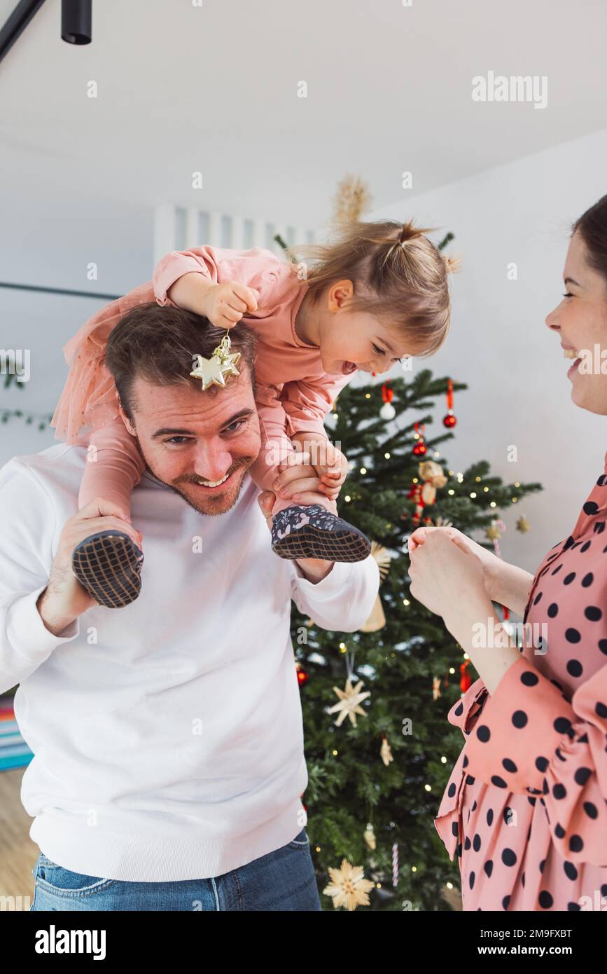 Dad carrying little girl on his shoulders smiling, while mom stands at the side looking at them - Family having fun on Christmas  Stock Photo