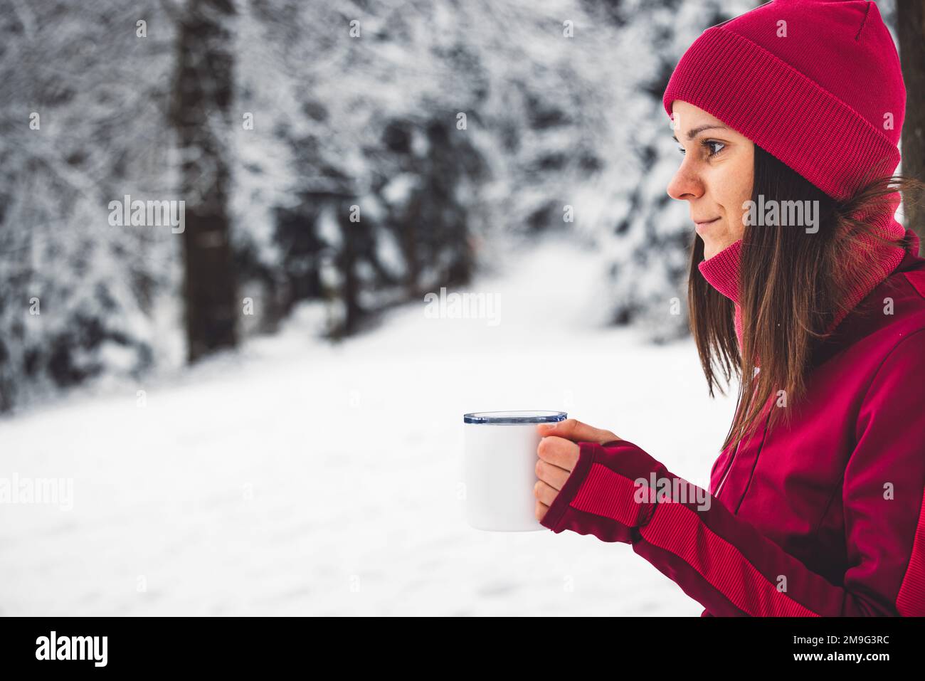 Side profile, waist up, woman in red jacket and hat drinking hot tea in the middle of a snowy winter forest Stock Photo