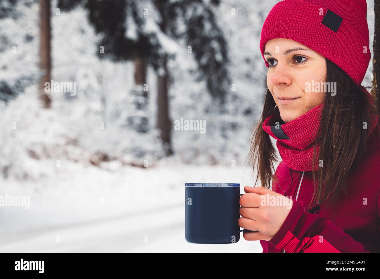 Portrait of caucasian woman in red winter outfit holding a blue metal reusable cup with tea in it - Winter forest Stock Photo