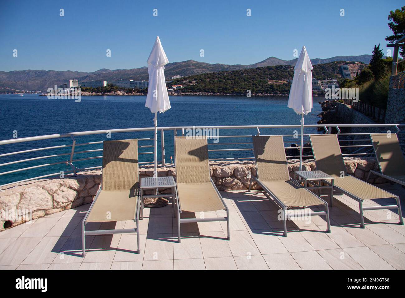 Empty sun loungers in a row at recreation area by the blue sea and a swimming pool. With lowered white parasols and blue sky. Dubrovnik, Croatia - Jul Stock Photo