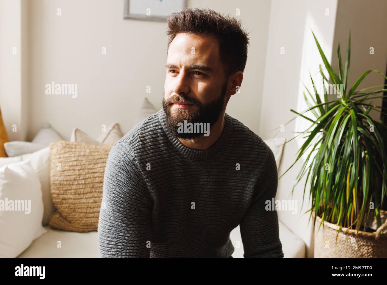 Thoughtful bearded biracial young man looking away while sitting on sofa in living room, copy space Stock Photo