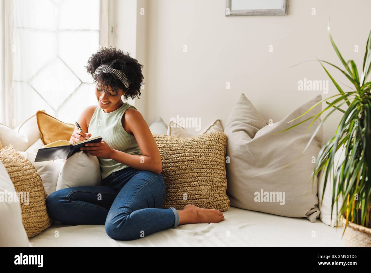 Smiling biracial young woman with afro hair writing in diary while sitting by window on sofa Stock Photo