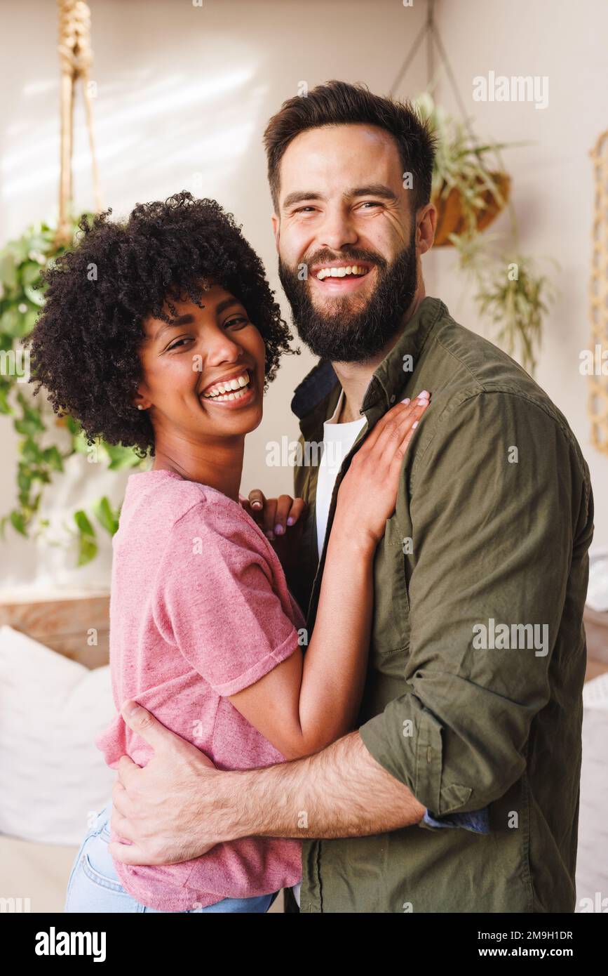 Portrait of cheerful biracial young couple laughing and hugging while standing against white wall Stock Photo