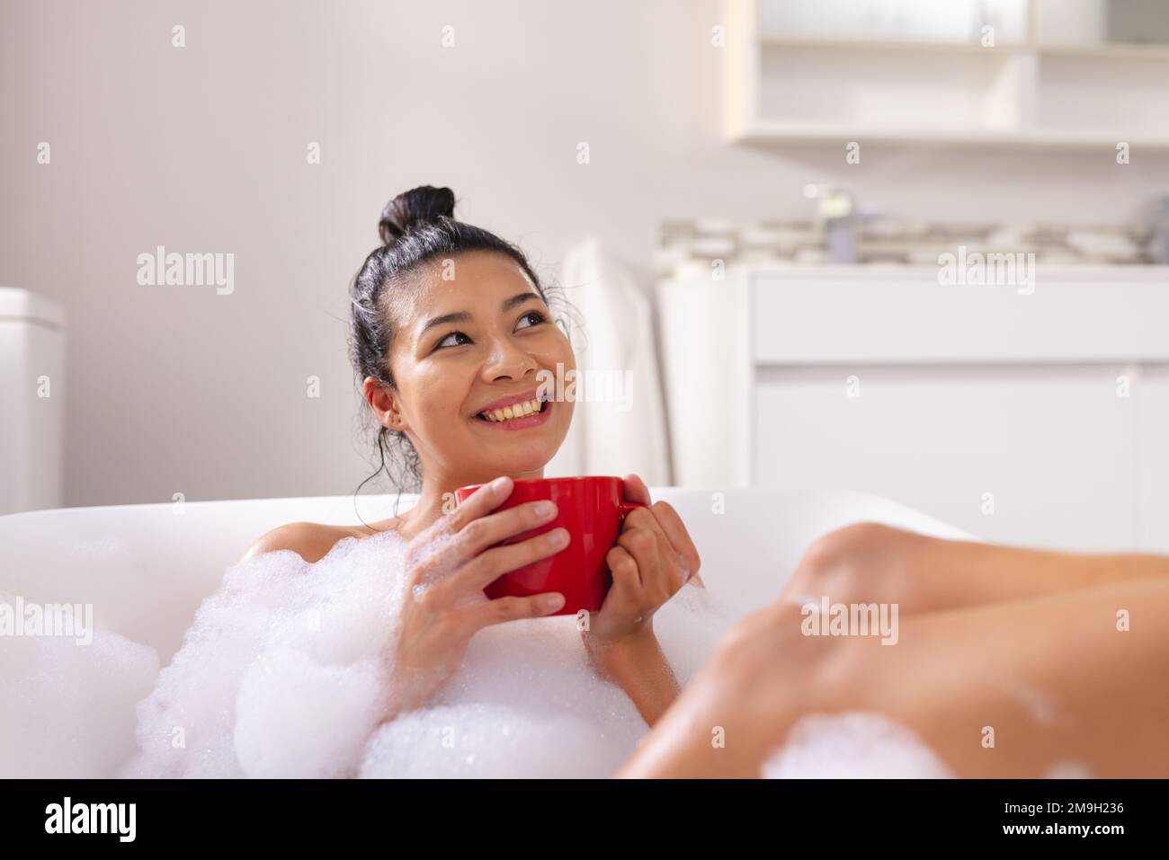 Image of biracial woman relaxing with cup of tea in bathtub in bubble