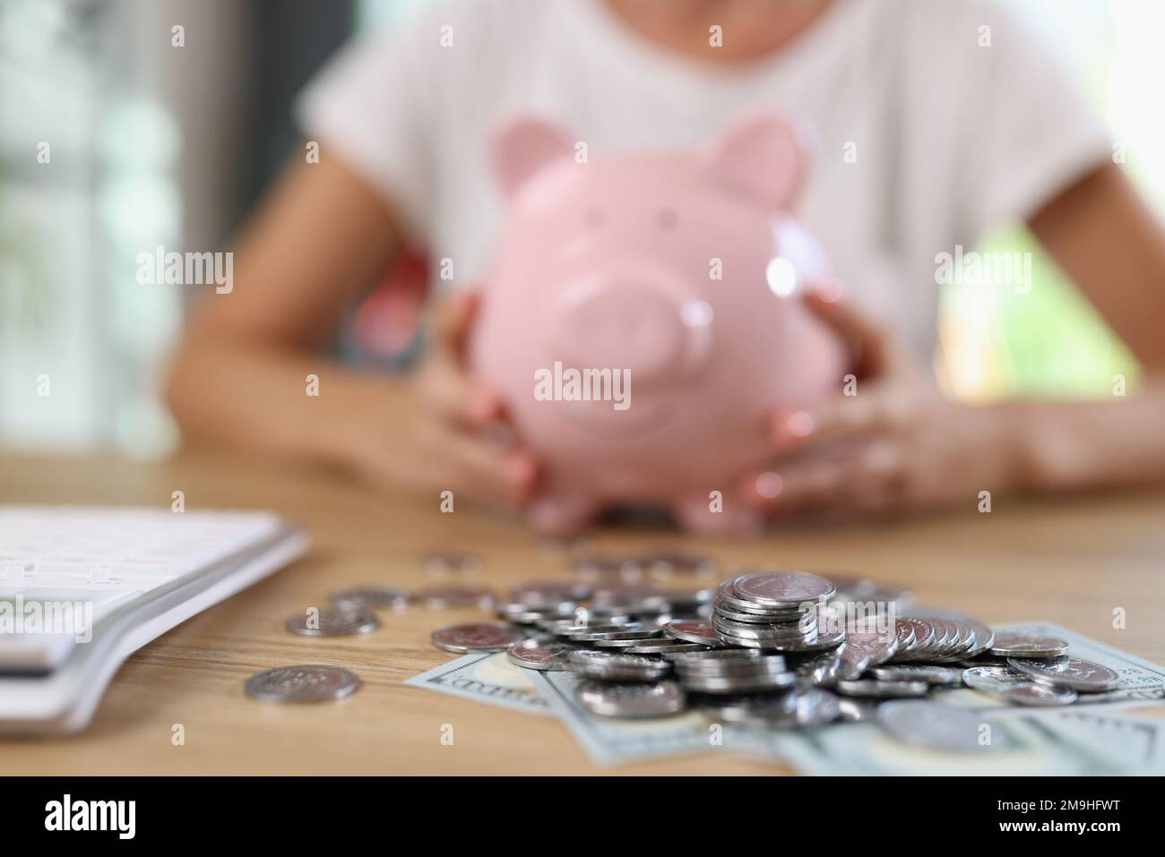 Banknotes and coins on table and blurry woman with piggy bank in background. Stock Photo