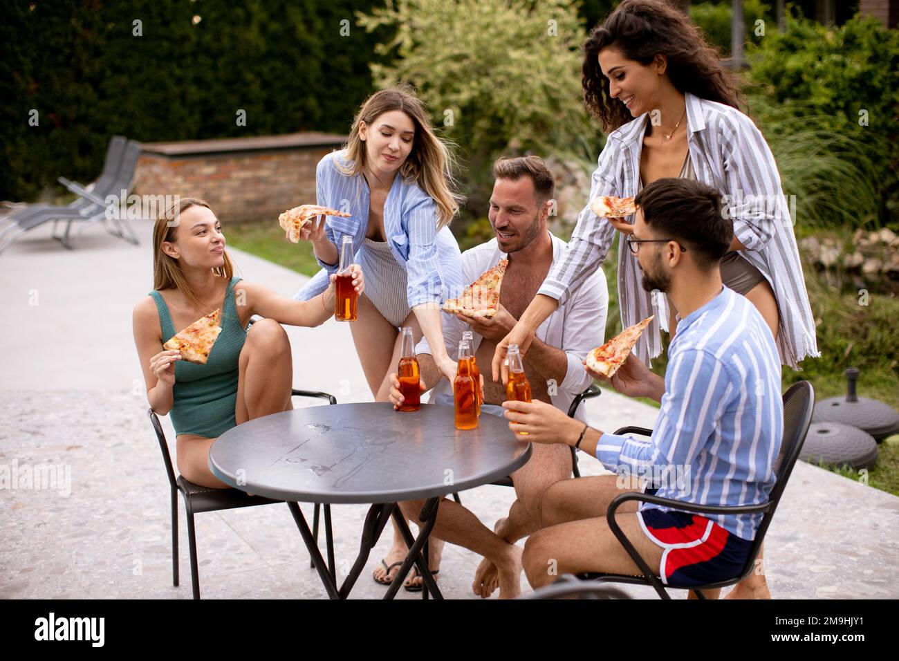 Group of happy young people cheering with cider and eating pizza by the pool in the garden Stock Photo