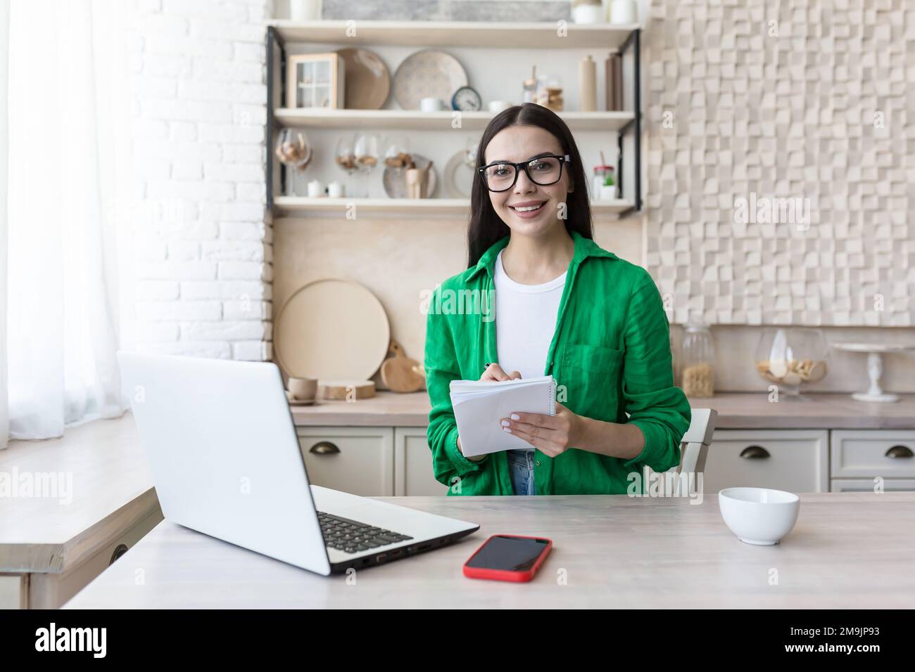 Portrait of a young woman teacher in glasses sitting at home in the kitchen with laptop. Works from home online. He is holding a notebook with a pen in his hands, smiling at the camera. Stock Photo