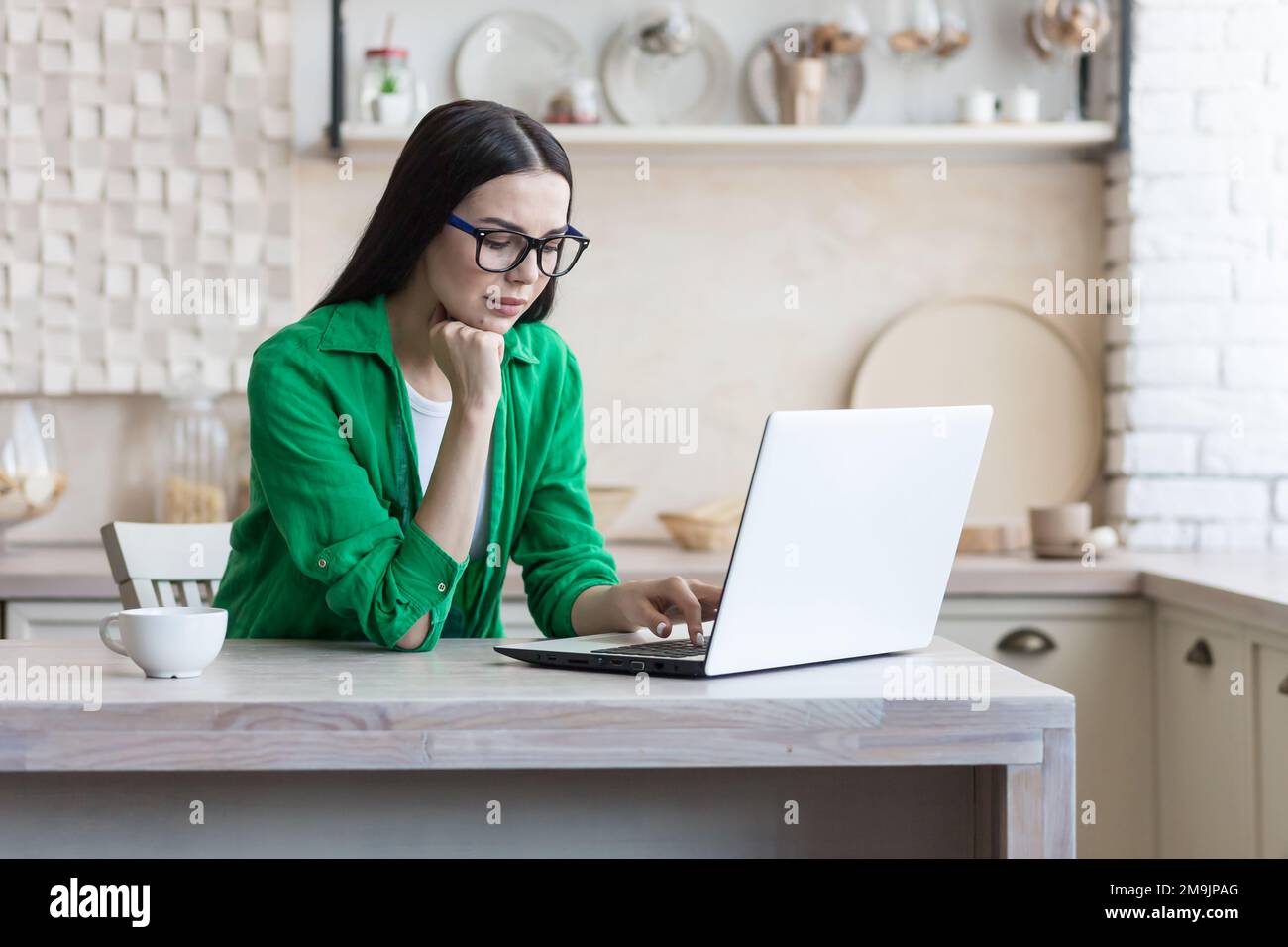Focused young female designer, engineer, architect working from home online. Sitting in the kitchen at the table, working on a laptop. He holds his head with his hand, looks at the monitor. Stock Photo