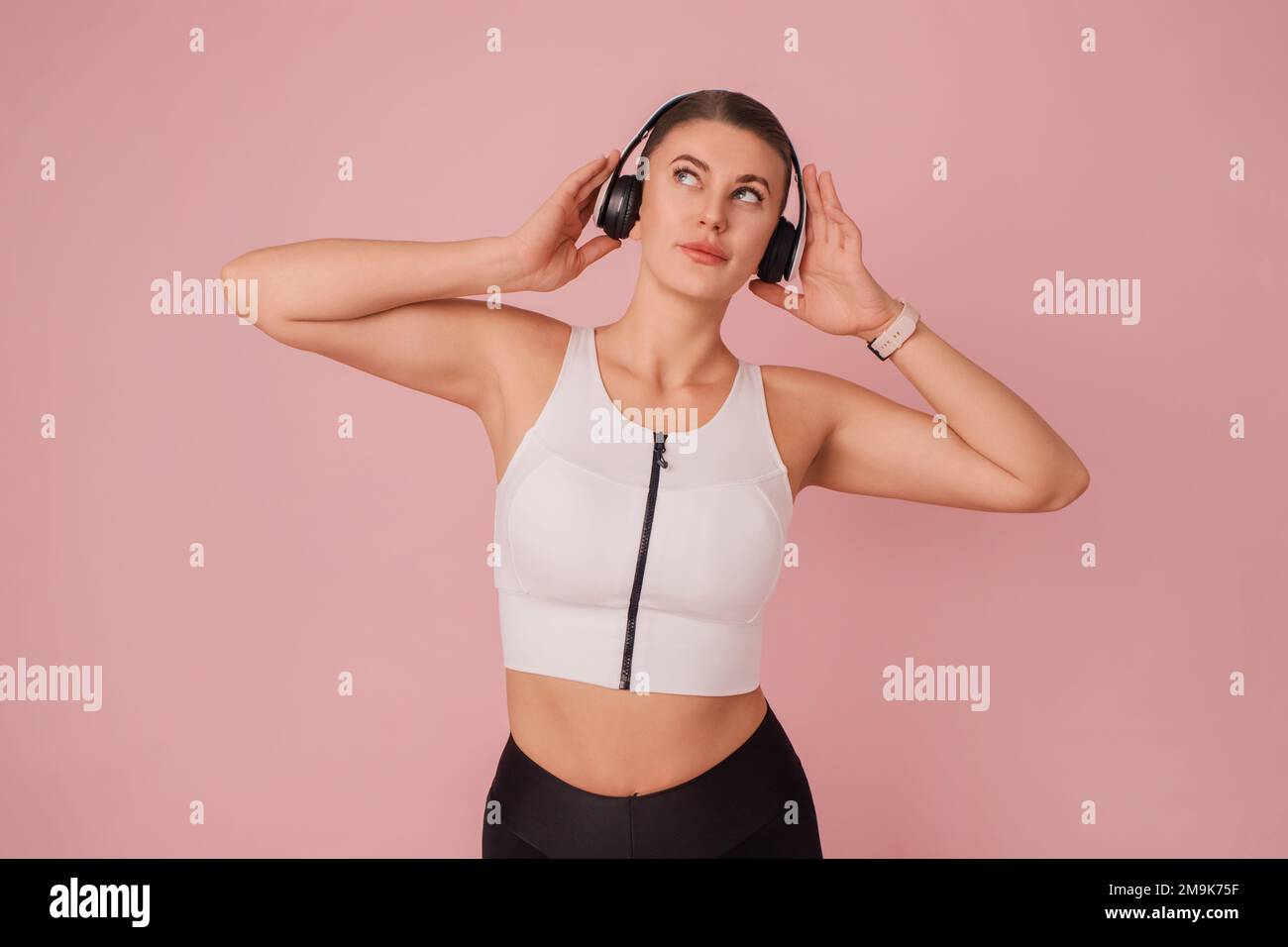 Stylish young woman listening music from white headphones. Girl in sportswear holding earphones and looking up on pink background. Studio portrait Stock Photo