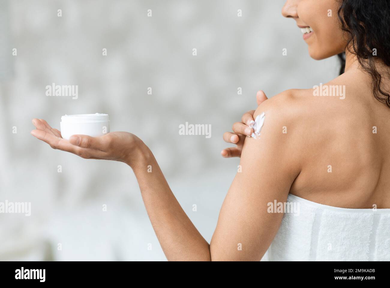 Smiling middle eastern millennial curly woman in towel holding jar, applies cream on her shoulder in bedroom Stock Photo