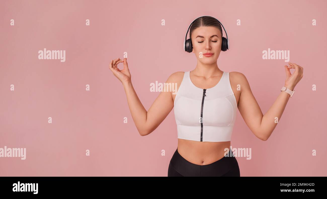 Meditation practice. Attractive girl with yoga gesture on pink backdrop. Portrait of young fitness woman listening relaxing sounds with closed eyes Stock Photo