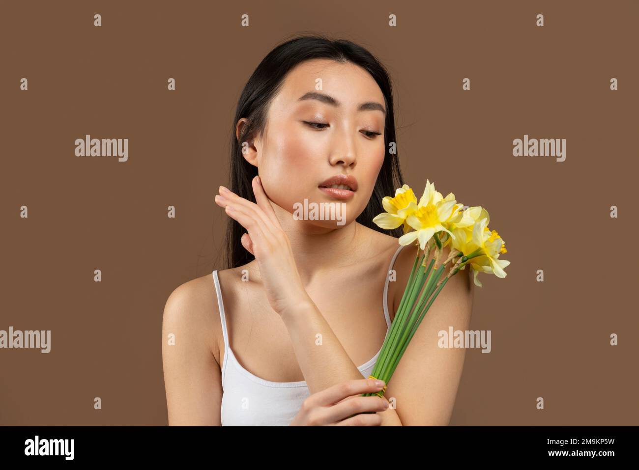 Young beautiful japanese lady posing with yellow daffodil flowers isolated on brown background, studio shot Stock Photo