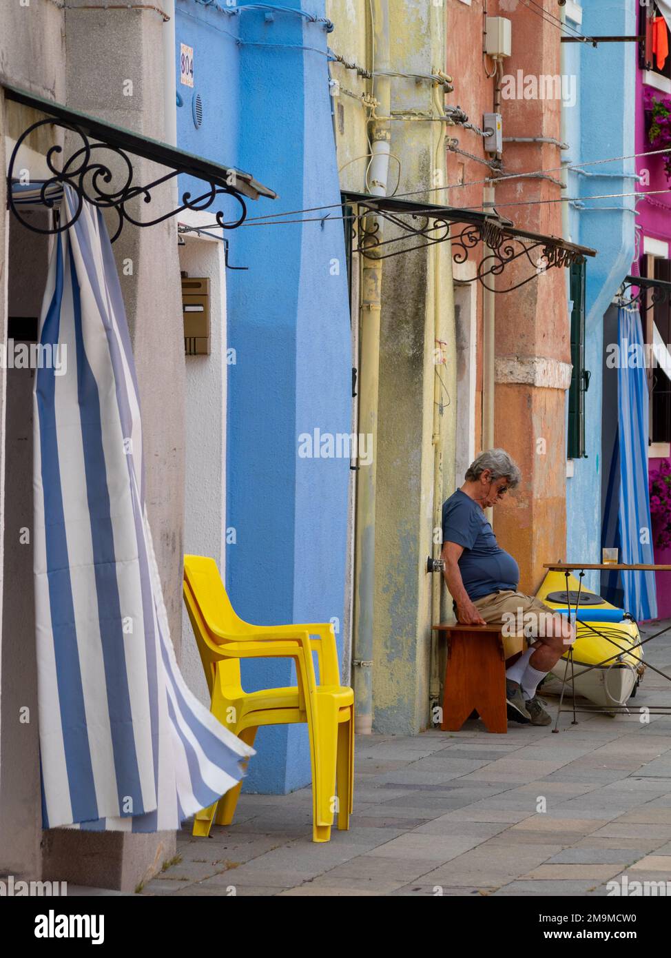 A man sleeps seated outside in a colorful street of Burano Stock Photo