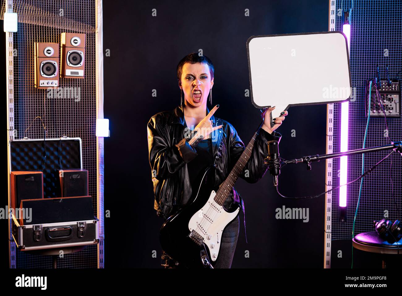 Musician showing blank placard with copy space for advertisement in music studio, person playing guitar preparing for rock concert. Rebel woman with leather outfit performing heavy metal song Stock Photo